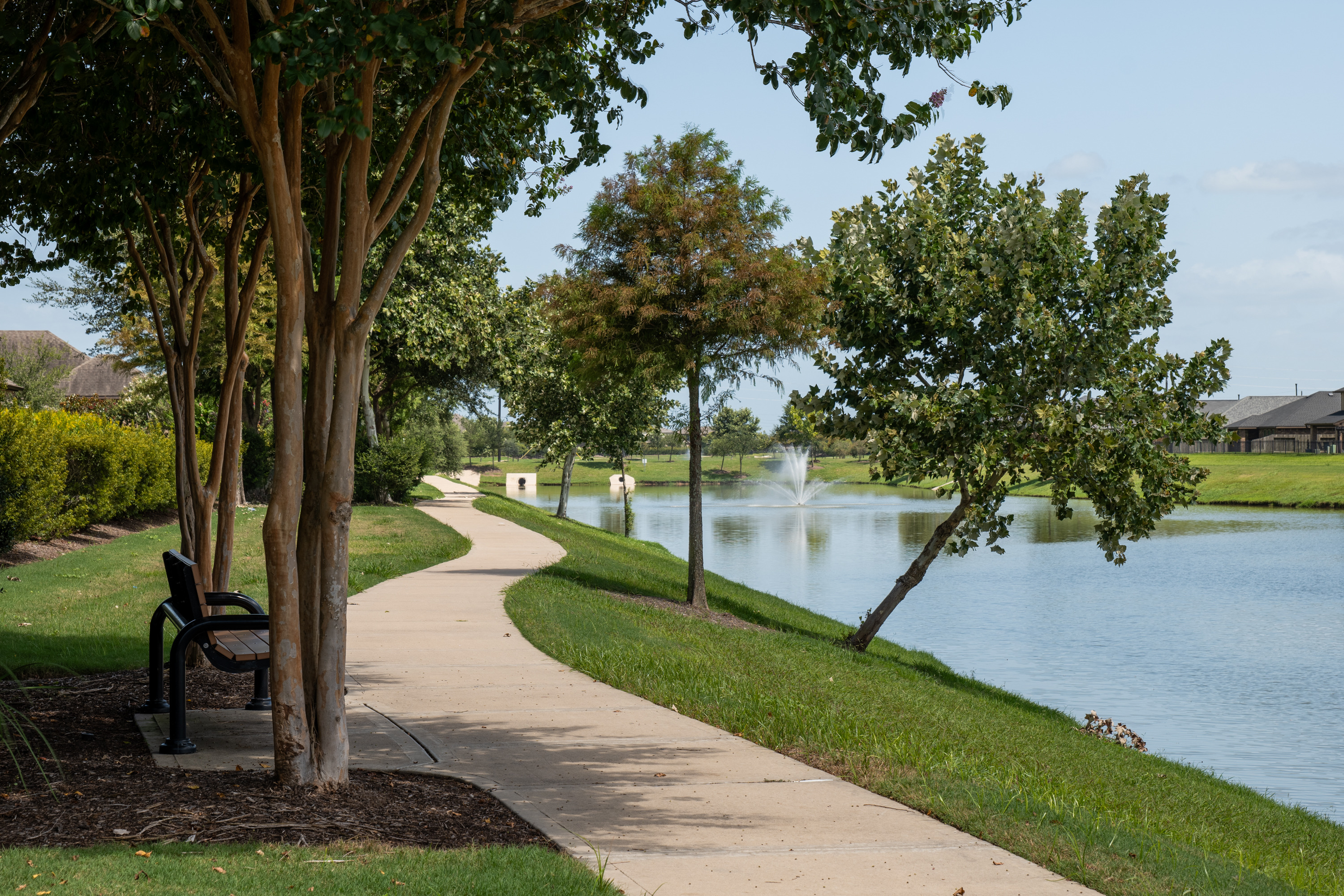 A shaded bench overlooks one of Bonbrook's seven lakes, with a paved trail running along the shore.